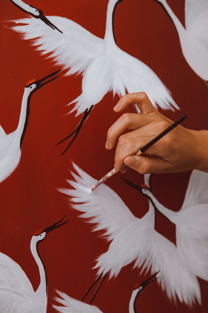 Close-up of a hand painting white cranes on a vibrant red canvas.